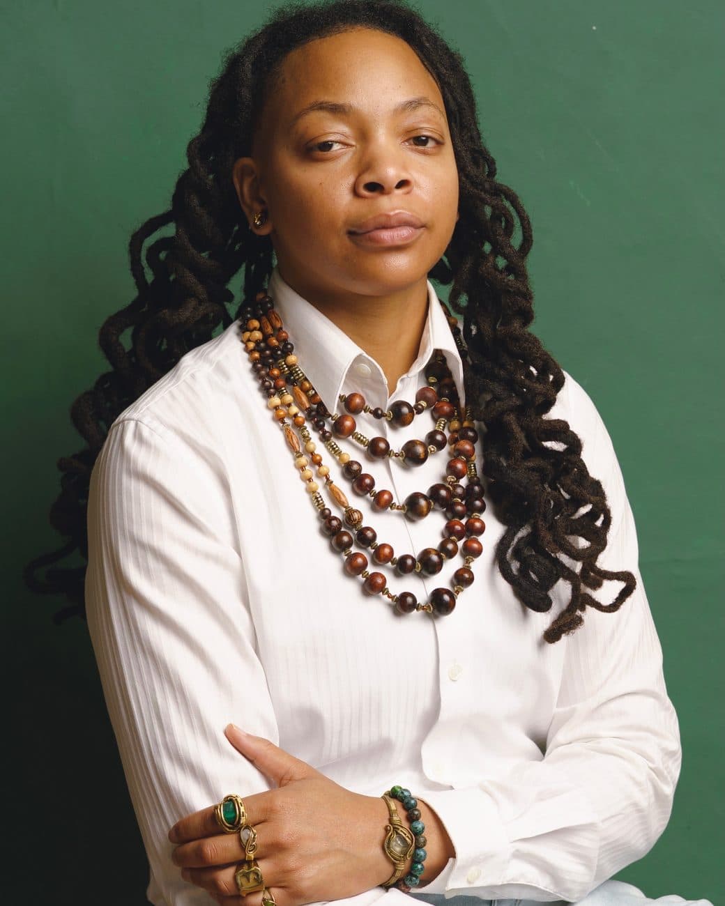 Portrait of Crys Adams in a white shirt with layered wooden-bead necklaces and brass rings, arms folded, against a green backdrop.