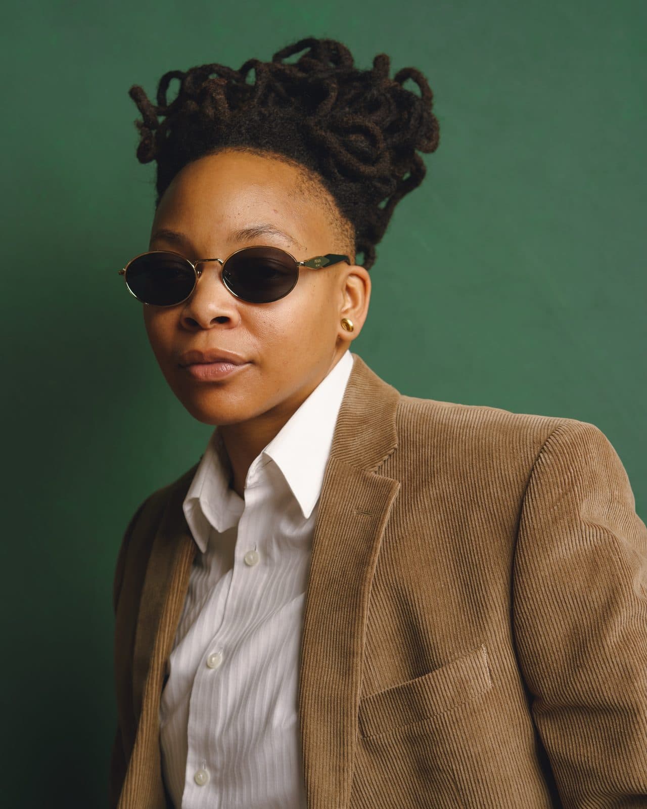 Portrait of artist Crys Adams against a deep green backdrop, wearing oval sunglasses and a camel corduroy blazer over a white shirt.