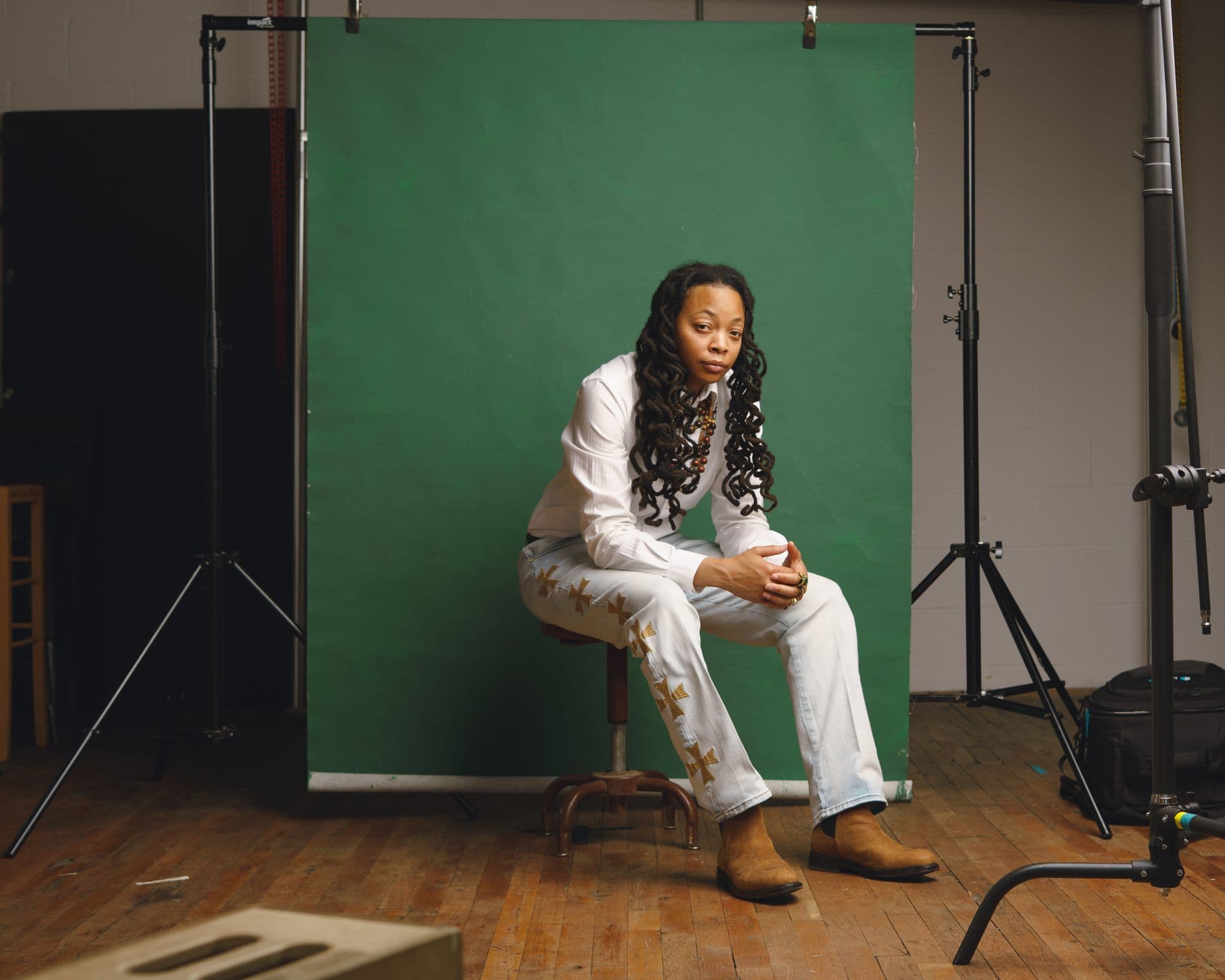 Crys Adams seated in her studio on a stool before a green backdrop, surrounded by lighting equipment.